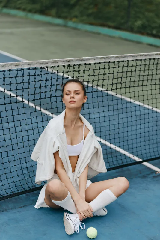 Young female athlete in stylish sporty attire, sitting on a tennis court, exuding confidence and strength, surrounded by vibrant colors of the court and net