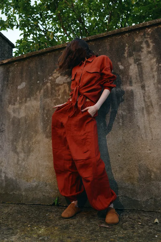 A stylish woman wears a loose red jumpsuit while leaning against a weathered wall, hands in pockets. The casual outdoor shoot highlights contemporary fashion and relaxed confidence