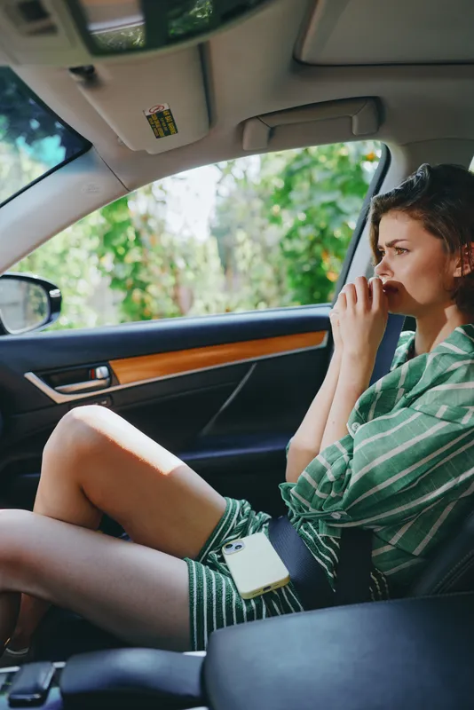 Casual man sits in a car interior, talking on a mobile phone, relaxed pose with leg up, natural daylight, green striped shirt, wooden trim accents.