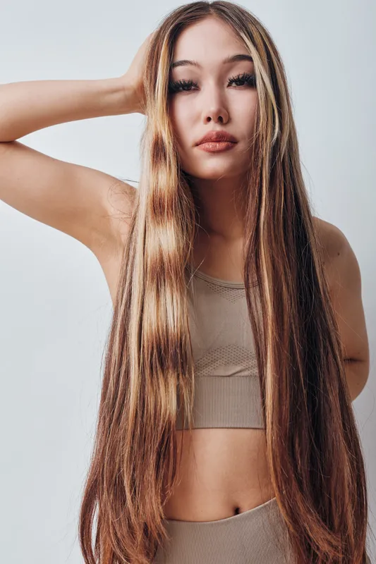 Woman with long straight hair posing in sleeveless top on plain background. Closeup portrait of female model with natural makeup and hand near head, looking at camera confidently.