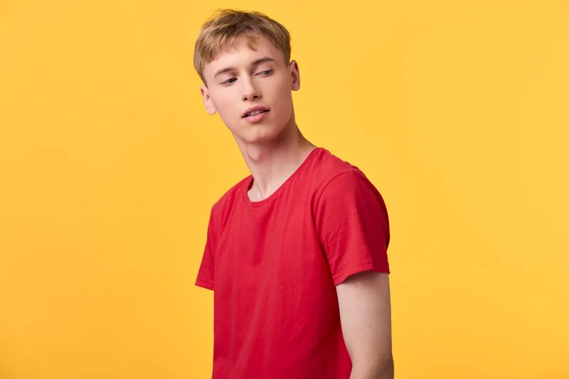 Young man in a red shirt stands before a vibrant yellow background, creating a bold fashion statement with a casual, relaxed pose and soft lighting that highlights modern street style.