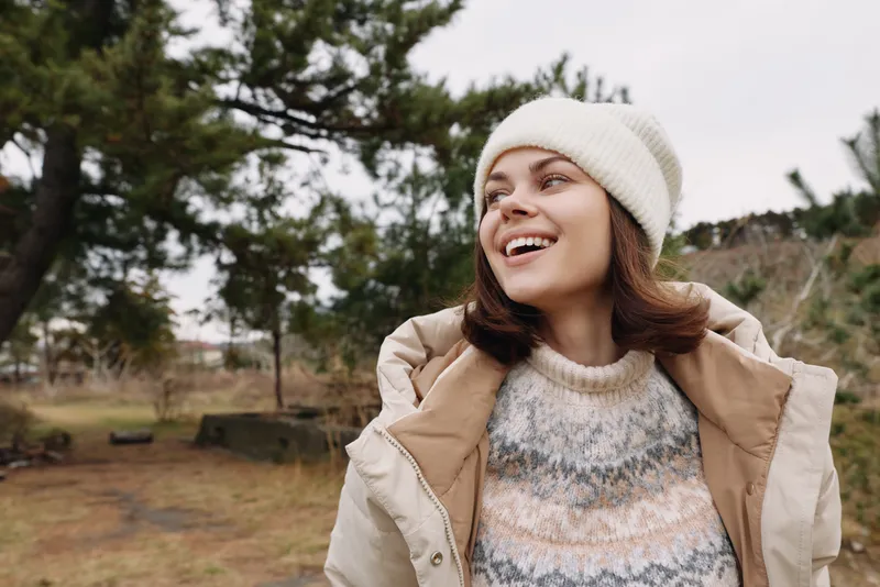 Smiling woman in a warm winter outfit outdoors, wearing a knit sweater, coat and beanie, enjoying fresh air in a park with trees, open space and natural light, candid lifestyle moment