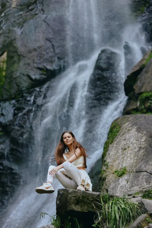 young woman sitting on rock near waterfall in nature wearing white outfit with casual shoes, relaxed pose, beautiful scenic outdoor background, natural light
