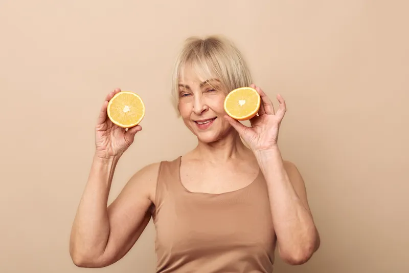 Smiling mature woman with short blonde hair holds two halves of fresh orange near face, eyes closed. Concept of healthy lifestyle, natural skincare, vitamin C, and wellness at home.