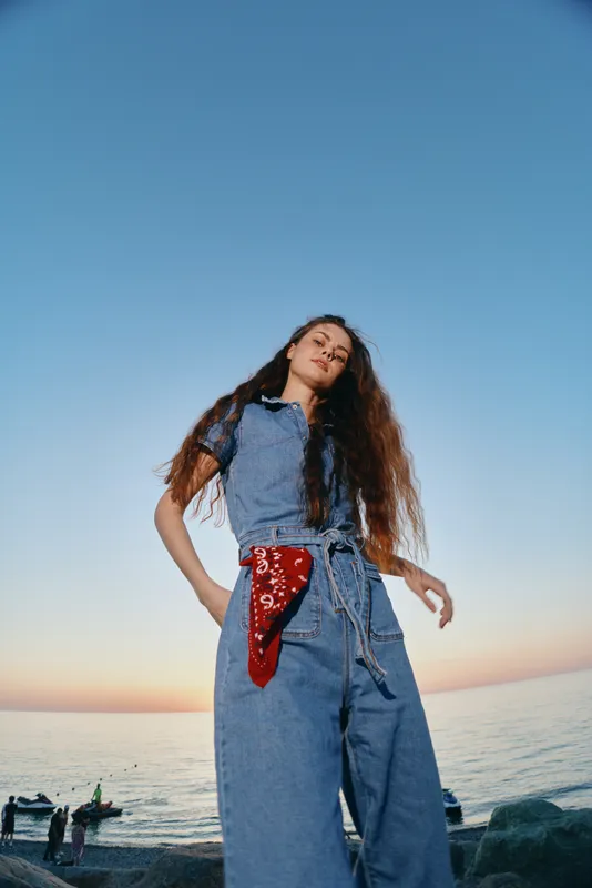 lifestyle woman in modern boho-western denim street style stands outdoors with sea and sunset background, film color tones create warm and nostalgic mood
