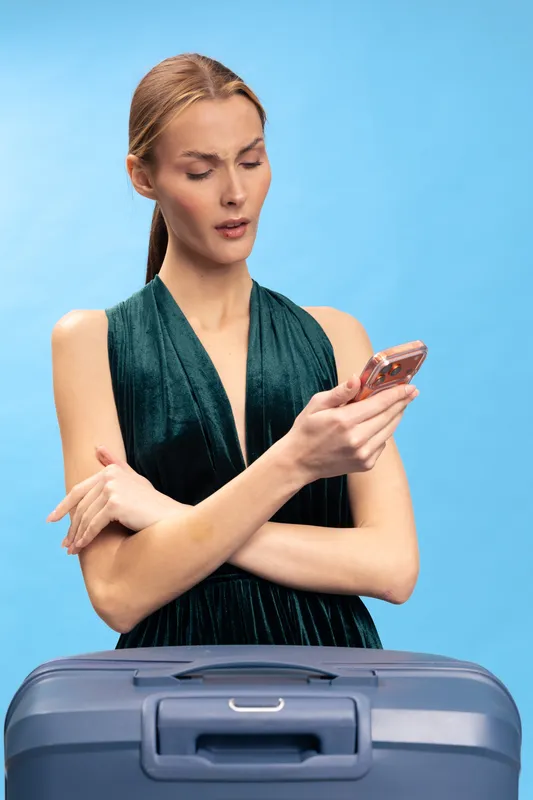 Confused woman in green dress looks at smartphone while standing behind blue suitcase. Concept of travel planning, uncertainty, and holiday preparation on bright blue background.