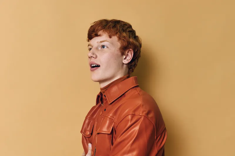 Young man with red hair wearing a leather jacket posing with emotional expression on beige background. Casual style, side view, isolated portrait in studio lighting.