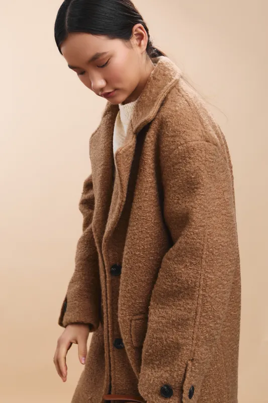 Asian woman in a warm brown coat stands in a soft studio setting, exuding calm and focus as she adjusts her sleeves and gazes downward.