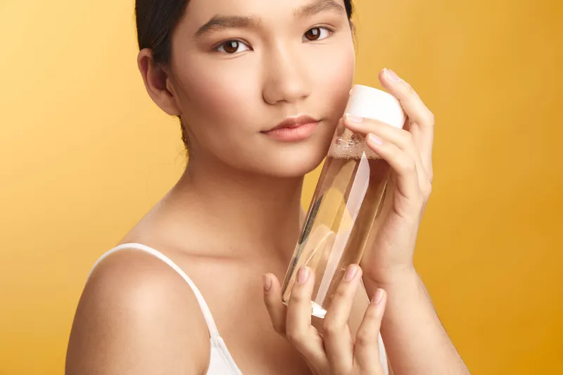 Asian woman holds a skincare product close to her face, posing against a warm backdrop. She appears calm and confident, conveying beauty care and personal wellness in a stylish studio setting.