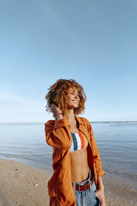 Young woman with curly hair enjoys a sunny day on the beach wearing a colorful top and orange shirt.