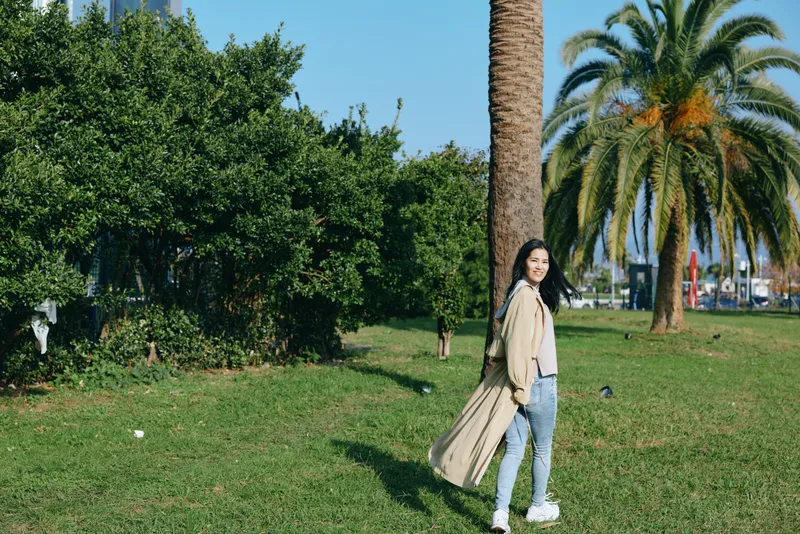 Woman walking in a sunny park with flowing coat, casual denim and bag, enjoying outdoor leisure among palm trees and green grass, fashion lifestyle scene