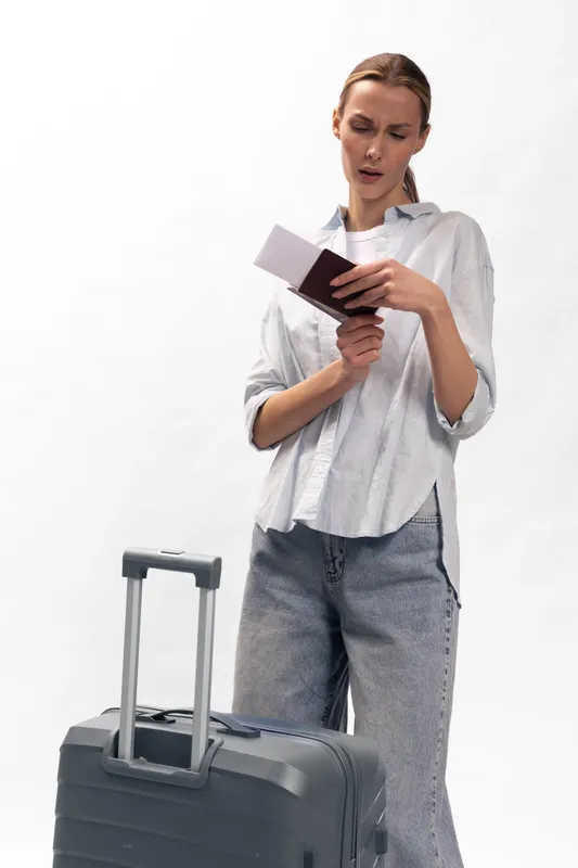 Young woman traveler in casual clothes checks passport and ticket while standing near suitcase with extended handle on white background. Concept of travel, trip preparation, and holiday planning.