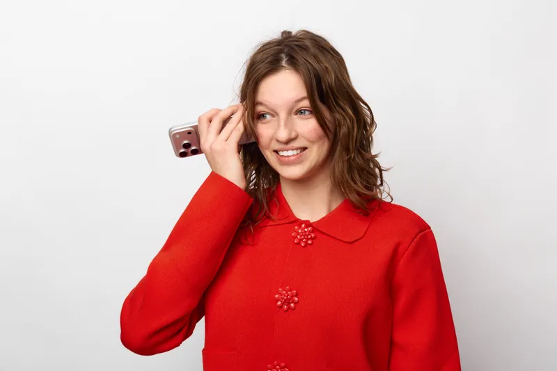Smiling young woman with curly hair wearing red sweater holds smartphone near ear, looking aside. Concept of communication, technology, casual lifestyle, and positive emotions in studio.