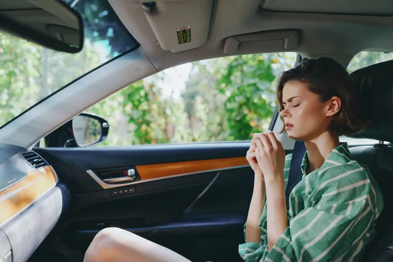 Woman sits inside a car applying makeup with a calm, focused expression, sunlight softly filtering through the window.