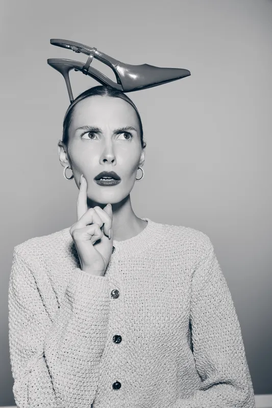 Young Caucasian woman with pale skin and short hair wears high heel shoe on head, looks confused. Studio portrait with grey background and casual textured sweater.