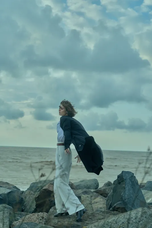 Woman standing on a rocky shoreline, wind catching her coat as the sea rolls in under a cloudy sky, fashion moment, solitary escape, graceful posture and calm reflection.