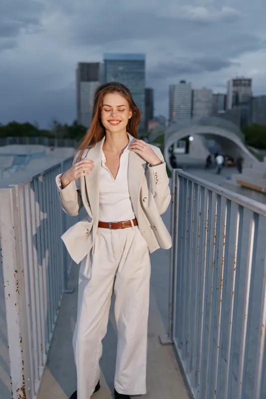 Young woman in beige business suit standing confidently outdoors with city skyline background smiling, urban fashion style, professional and optimistic mood
