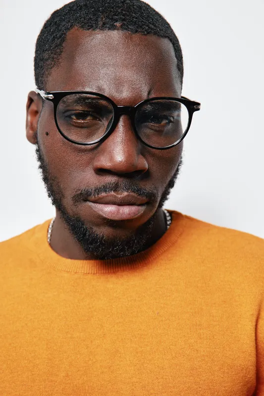 African American man with dark skin wearing black glasses and orange sweater looking serious on white background. Closeup portrait of thoughtful young male with facial hair and short hair.
