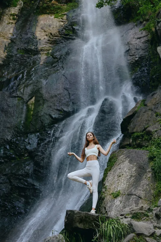 young woman meditating in white sportswear near a large waterfall, surrounded by green foliage and rocky cliffs, outdoors in natural light