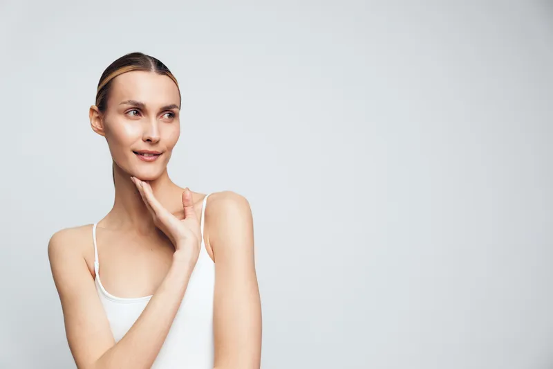 Portrait of a smiling young woman with clear skin wearing a white tank top, looking to the side on a plain light gray background, concept of beauty, health, and natural skincare.
