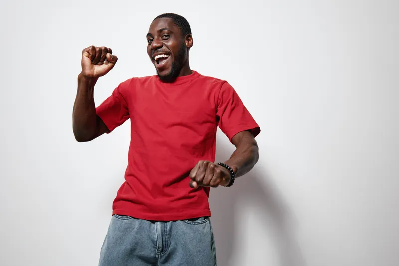African American man with dark skin wearing red t shirt and jeans dancing happily against white background with joyful expression and raised fists.