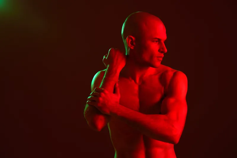 Athletic man posing in a dramatic red light. The image captures strength and boldness, showcasing the model's muscular physique in a striking manner.