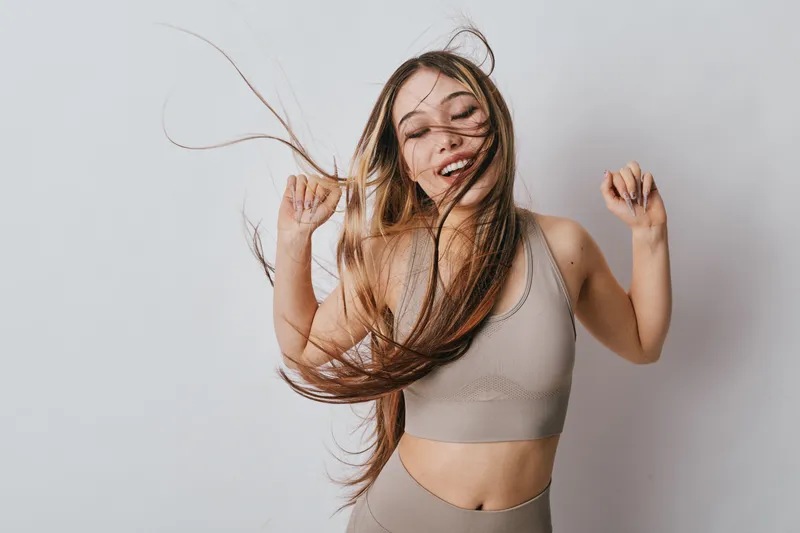 Young woman with long hair wearing sportswear dancing with eyes closed in studio. Healthy lifestyle female smiling with raised hands in beige fitness outfit and flowing hair motion.