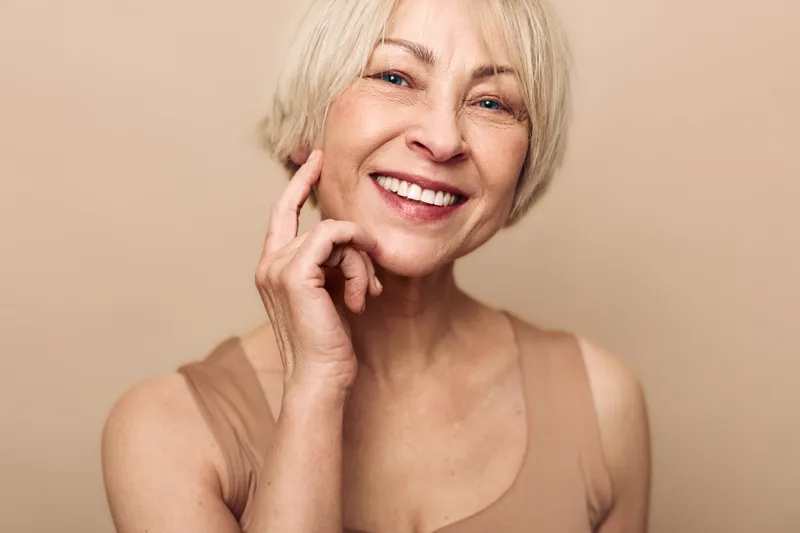 Smiling mature woman with short blonde hair touches her face gently, expressing confidence and natural beauty on beige background. Concept of aging gracefully and self-care.