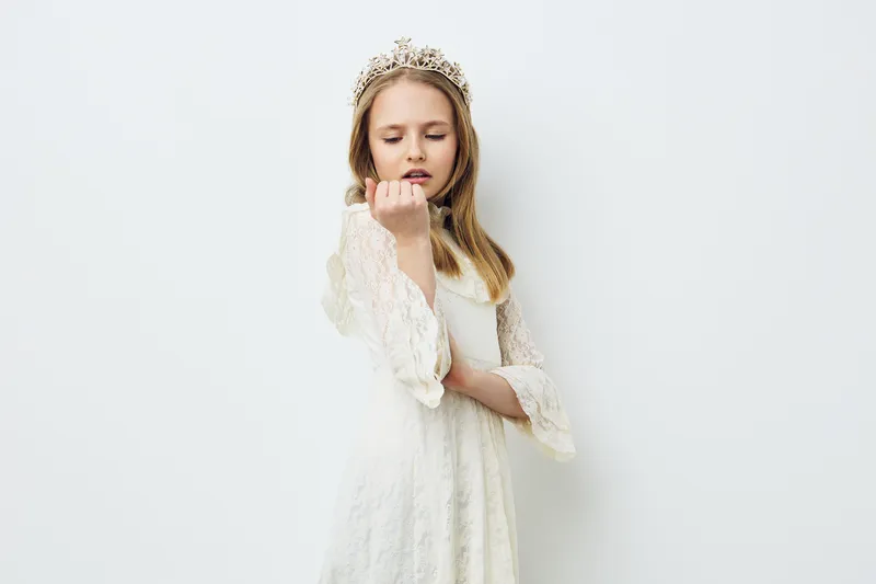 A young girl in a white dress with a crown poses thoughtfully against a minimalist background, conveying innocence and a touch of royalty