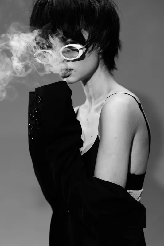 Black and white portrait of a young woman exhaling smoke wearing sunglasses and a loose jacket with a moody atmosphere.