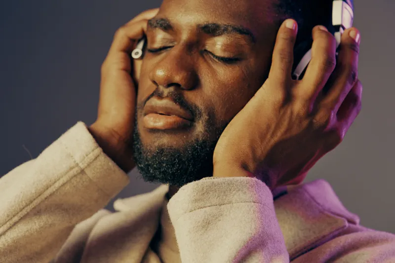 A young Black man enjoying music with headphones, deeply immersed in sound, showcasing emotions of relaxation and joy against a soft, colorful background