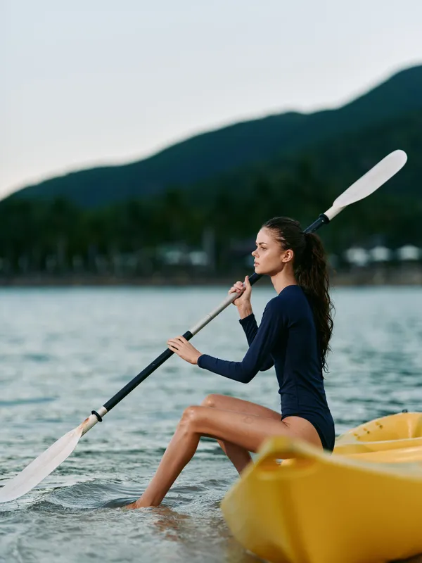 Woman paddling a yellow kayak on calm water near shore, raising a paddle mid stroke. Focused posture and serene landscape create a peaceful outdoor sports moment. The scene emphasizes balance,
