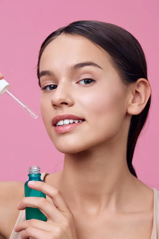 Young woman applying facial serum with a dropper for skincare on a pink background, showcasing beauty and health