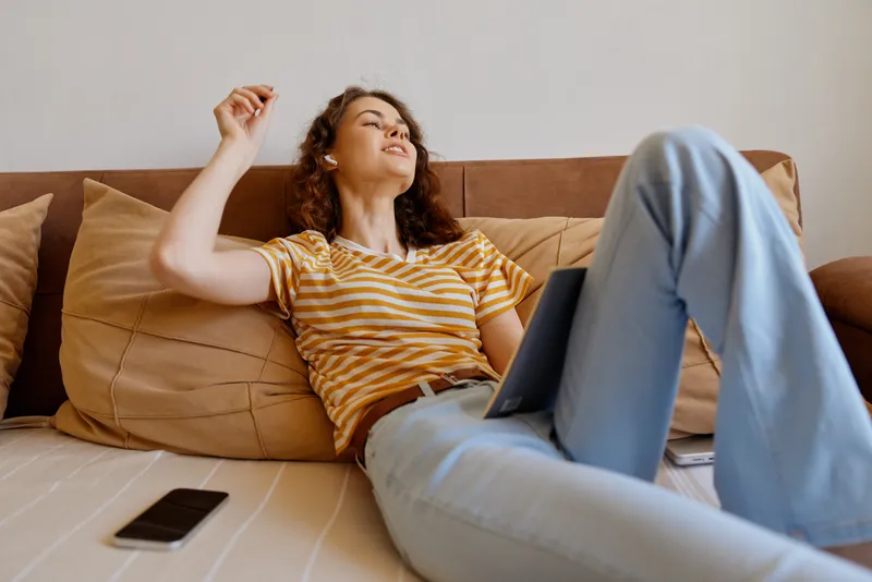 A woman relaxing on a couch at home with a laptop, enjoying casual comfort in a bright living space. A calm, everyday moment highlighting lifestyle and home relaxation.