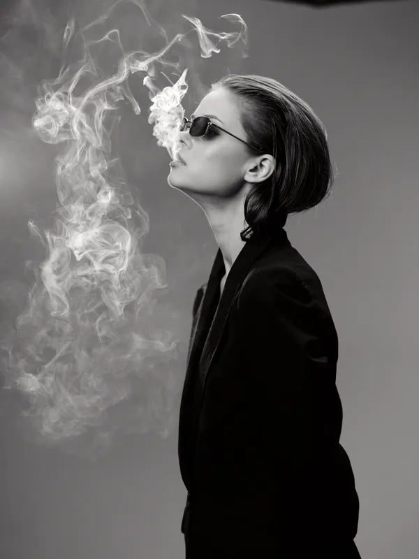 Black and white portrait of smoking woman in sunglasses with smoke swirling around her face in stylish outfit