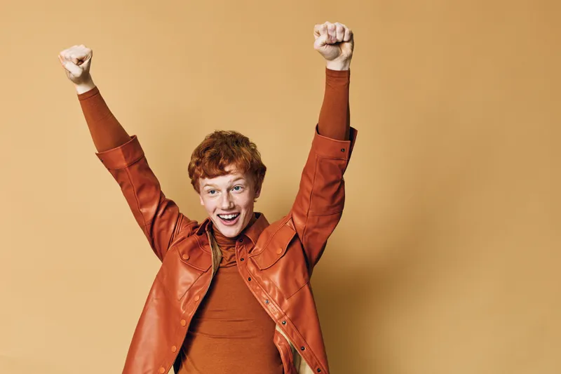 Young man with red hair raising fists and smiling with excitement against beige background. Casual clothing, energetic gesture and happy expression for success or celebration.