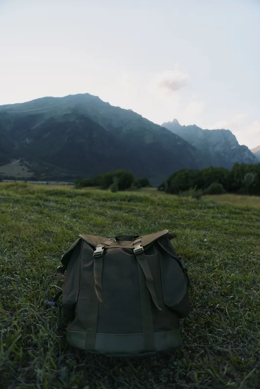 A green backpack stands alone on grassy ground with mountains in the background under a clear sky during early morning light.