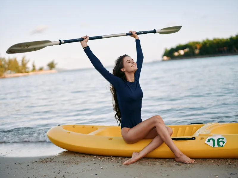 Smiling woman raising kayak paddle sitting on yellow kayak by the beach, enjoying sunny day, water and nature calmness near shore.