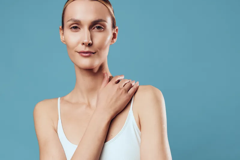 Portrait of calm woman with natural skin, wearing white tank top, posing with hand on shoulder, minimal makeup, studio lighting, plain blue background, serene expression