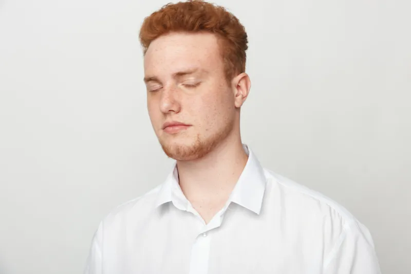 Young man with red hair and beard wearing white shirt closes eyes in calm expression. Isolated on white background. Concept of relaxation, peace, and mindfulness in portrait photography.