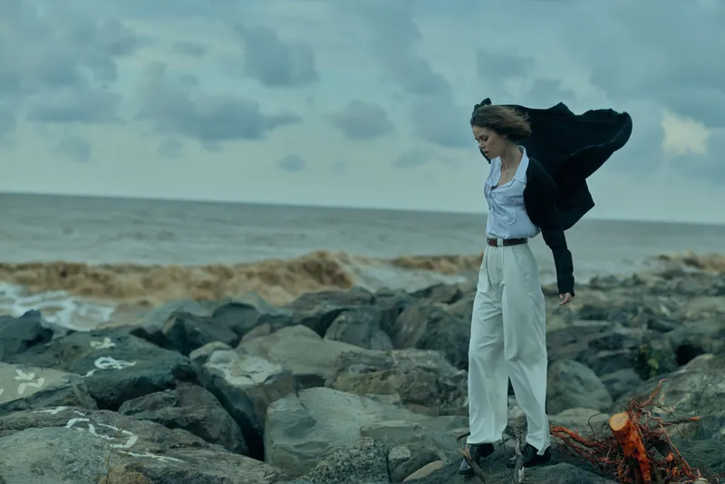 Female on a rocky shoreline facing strong wind with flowing coat, casual chic fashion scene by the sea, capturing motion, balance and resilience against the ocean backdrop.