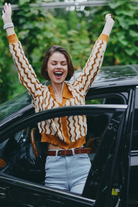 Young woman celebrates with arms raised from an open car door, capturing a bright, spontaneous moment outdoors with vibrant fashion.