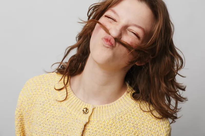 Young woman with curly brown hair makes a playful mustache with her hair, smiling with closed eyes. She wears a yellow knitted sweater against a plain light background. Fun and joy concept.