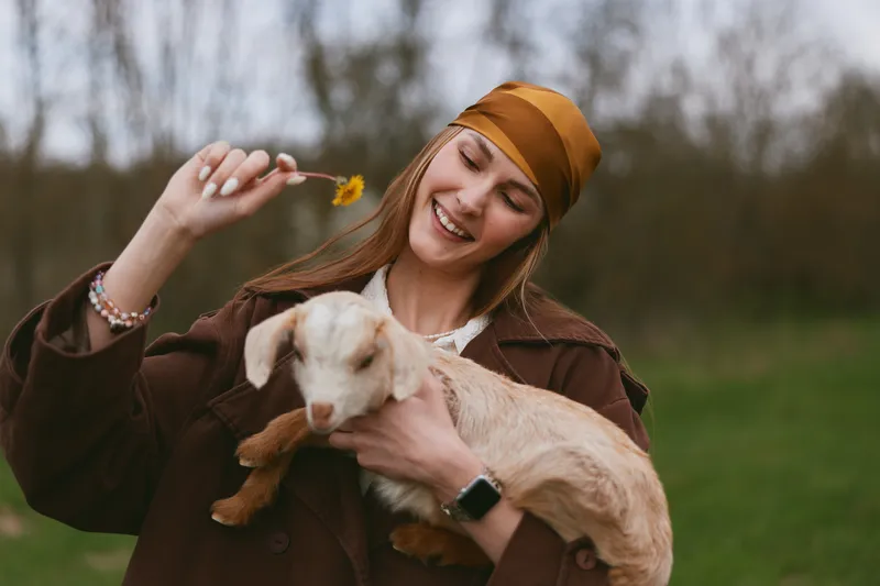A joyful woman in a brown jacket and mustard headscarf holds a small dog, smiling as she offers a yellow flower. An outdoors lifestyle moment capturing warmth and simple affection.