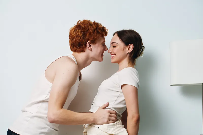 Portrait of smiling young couple embracing and leaning towards each other in white casual clothes indoors with lamp on white wall background, expressing affection and joy.