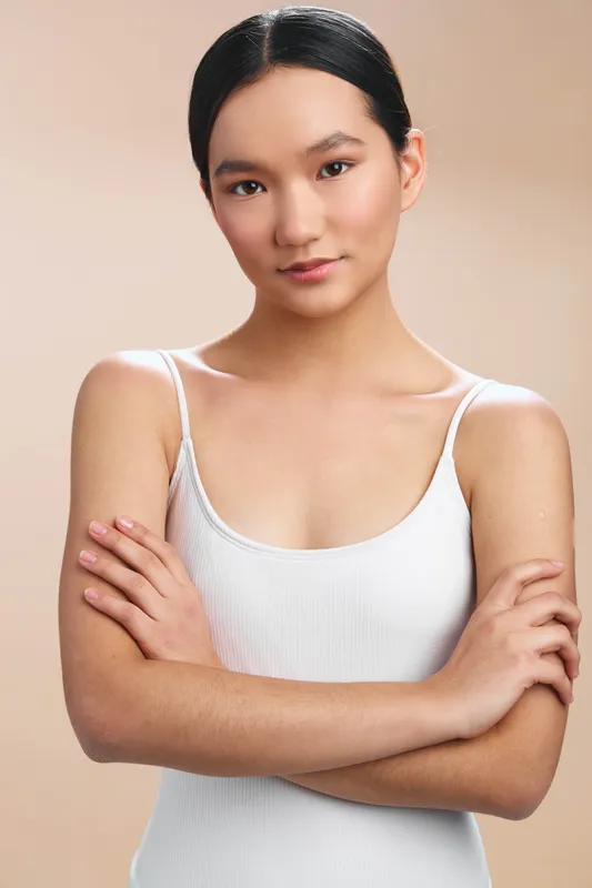 Asian woman stands in a neutral studio backdrop, arms crossed, calm expression, stylish white tank top, conveying confidence and poise with a modern, minimal vibe.
