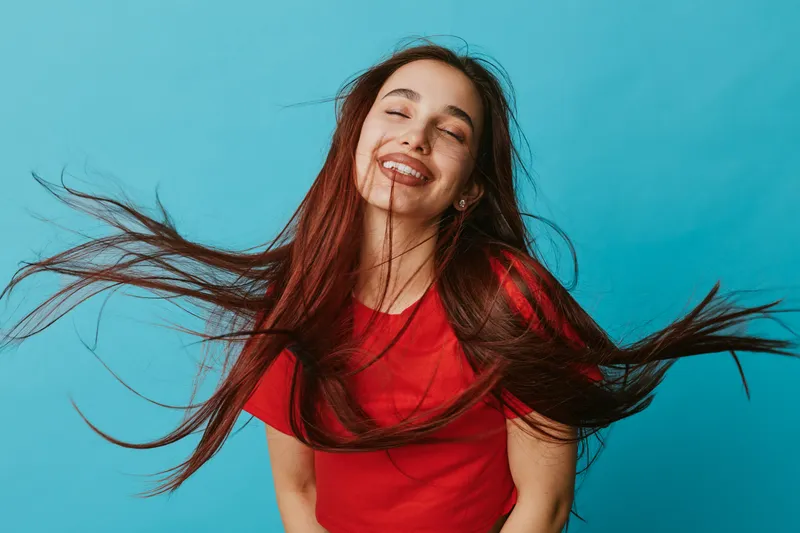 Young woman with long hair wearing red shirt smiling with closed eyes against blue background. Hair flying in motion, happy female portrait with natural expression and joyful mood.
