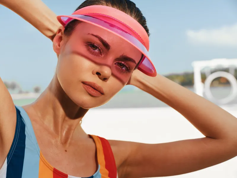 Close up portrait of a fashion model on a sunny beach, wearing a pink visor and colorful striped top, sunlit skin, calm expression, vibrant mood.