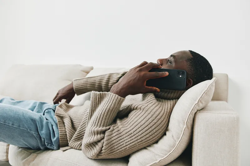 Relaxed young man in a cozy sweater holding a smartphone on a couch, exuding casual comfort and modern lifestyle vibes in a neutral toned living room