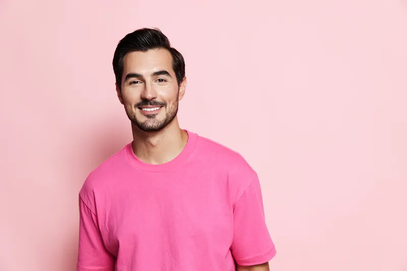 Smiling young man in a vibrant pink shirt stands confidently against a soft pink background, exuding a cheerful and approachable vibe.